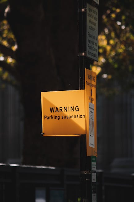 A close-up photograph of a yellow parking suspension warning sign mounted on a black metal pole outside a property in Harmondsworth, with additional smaller notices attached below it. The warning sign, visible in the foreground, clearly indicates parking restrictions for house removals or moving activities, which is relevant for planning vehicle access during furniture transport or home relocation. In the background, there are out-of-focus trees with green leaves and a grey fence, suggesting an outdoor environment and possibly a residential area. This image captures the typical signage used for scheduling and managing parking suspensions in relation to house removals, with natural lighting highlighting the details of the signage and pole. Man with Van Harmondsworth occasionally manages parking permits and suspension notices to facilitate a smooth loading process during furniture transport and packing and moving services.