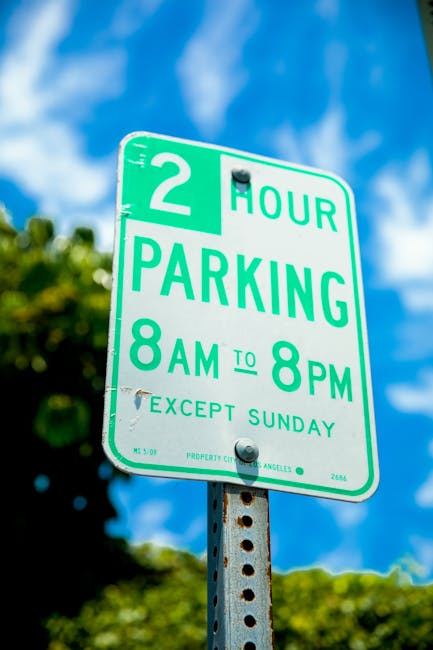 A green and white parking restriction sign mounted on a metal pole outdoors, indicating a two-hour parking limit from 8 am to 8 pm, Monday to Saturday, with no restrictions on Sundays, against a background of a blue sky with some clouds and green foliage. The sign is part of a street scene in Harmondsworth, associated with local parking regulations relevant to house removals and moving services offered by Man with Van Harmondsworth. The signage is clearly visible, and the environment suggests daytime in a residential or suburban area, where parking permits and suspensions may be necessary for furniture transport during home relocation or moving projects.