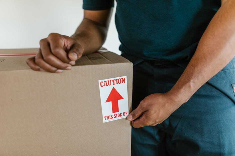 A person wearing a dark green t-shirt and navy blue trousers is seen securing a large cardboard box during a home relocation. The individual is gripping the box firmly with both hands, with one hand near the top and the other near the bottom. A red and white caution label on the box indicates 'CAUTION THIS SIDE UP' with an arrow pointing upwards. The box is placed on a flat surface indoors, possibly on the floor or a table. The background is neutral and uncluttered, emphasizing the focus on the handling of the moving box. This scene is part of a furniture transport or packing and moving process, which has been carried out by Man with Van Harmondsworth, a professional removals service specializing in household moves.