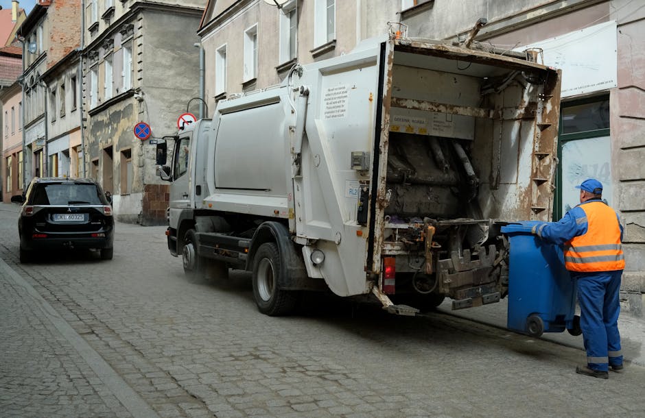 A street scene showing a waste collection vehicle operated by Man with Van Harmondsworth, positioned parallel to a row of old, multi-storey residential buildings with weathered facades and vandalized exterior walls. The truck, parked on cobbled pavement, has its rear hatch open, revealing an empty, rusty container designed for bulky waste pickup. A worker dressed in blue protective clothing, including a high-visibility orange vest and blue gloves, is standing beside the vehicle, emptying a blue wheeled bin into the collection hopper. The scene is lit by natural daylight, with a black parked car visible in the background. The environment reflects urban residential surroundings, supporting house removals, packing, and logistics activities related to home relocation services provided by Man with Van Harmondsworth, aligned with the context of bulky waste collection for residents in Harmondsworth Moor.