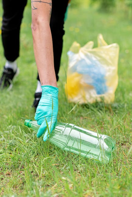 A person wearing a turquoise rubber glove is picking up a clear plastic bottle from a grassy outdoor area, with their arm extended downward. In the background, there is a large yellow plastic bag filled with other recyclable materials and some greenery. The scene is set on a bright day with natural daylight illuminating the grass and objects, reflecting the environmental aspect of waste disposal. This image depicts the process of environmental cleanup as part of house relocation or move-related duties, aligning with services provided by Man with Van Harmondsworth, such as packing, waste removal, and furniture transport during home relocation or move management.