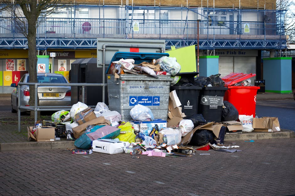 A street scene showing a waste collection vehicle operated by Man with Van Harmondsworth, positioned parallel to a row of old, multi-storey residential buildings with weathered facades and vandalized exterior walls. The truck, parked on cobbled pavement, has its rear hatch open, revealing an empty, rusty container designed for bulky waste pickup. A worker dressed in blue protective clothing, including a high-visibility orange vest and blue gloves, is standing beside the vehicle, emptying a blue wheeled bin into the collection hopper. The scene is lit by natural daylight, with a black parked car visible in the background. The environment reflects urban residential surroundings, supporting house removals, packing, and logistics activities related to home relocation services provided by Man with Van Harmondsworth, aligned with the context of bulky waste collection for residents in Harmondsworth Moor.