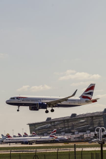 A British Airways commercial airplane is captured mid-air during descent at Heathrow Airport, with its landing gear extended and flaps deployed as it approaches the runway. The aircraft displays the British Airways livery with a predominantly white fuselage, the airline's logo, and the Union Jack design on the tailfin. In the background, part of the airport terminal building with large glass windows is visible, along with several other British Airways aircraft parked on the tarmac, their tails showing the airline's branding. The scene is set against a partly cloudy sky with a few scattered clouds, and the airport's perimeter fencing is seen at the bottom of the image. This scene illustrates the busy environment typical of home relocation and furniture transport logistics, where efficient aircraft movement is essential for timely moving services coordinated by companies like Man with Van Harmondsworth, particularly in the context of Heathrow area moves and packing and moving operations.