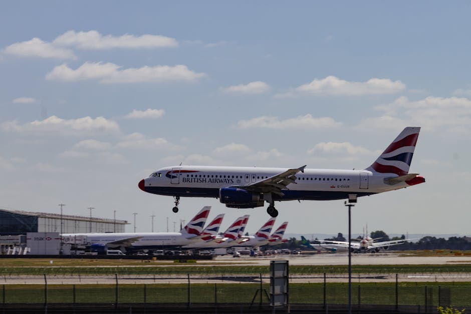 A British Airways aircraft is captured mid-air, descending towards a runway at an airport during the daytime. The airplane's fuselage displays the British Airways logo and branding, with the aircraft's landing gear extended as it prepares to land. In the background, multiple British Airways planes are parked on the tarmac, with their distinctive tail fins featuring the airline's red, white, and blue stripe design. The scene includes airport terminal buildings, runway markings, and a partly cloudy sky overhead, suggesting clear weather conditions. This image visually illustrates air travel and the logistical process of transporting passengers and goods efficiently, aligning with contents about house removals, packing, and moving services by companies like Man with Van Harmondsworth, supporting seamless relocation logistics in the Heathrow area.