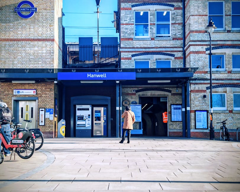 The image captures the exterior entrance of Hanwell station, which features a brick building with a blue sign displaying the station name. In front of the station, there is a paved pedestrian area with a person wearing an orange coat and dark trousers standing and looking towards the entrance. To the left, several bicycles, including a prominent red and black bike, are parked near bike racks. Visible on the left side of the building are ticket vending machines, an information display panel, and a small lift station for accessibility. The station's entrance includes an archway leading into the underground platform, and an orange safety vest worn by a staff member is partially visible through the arch. The environment is well-lit with daylight, and some railings and lampposts are seen along the pavement, creating an urban setting suitable for housing relocations or moving logistics planning, consistent with services provided by Man with Van Harmondsworth.