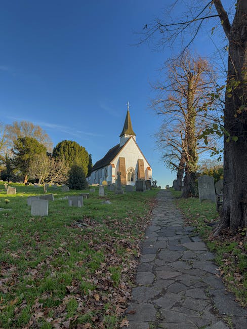 A narrow, uneven stone pathway leading up a grassy slope toward a small white church with a tall, pointed steeple, situated among trees and old gravestones in a churchyard under a clear blue sky; the scene captures the quiet environment of a rural or historical area during daylight, with visible shadows cast by the trees and the church, illustrating the outdoor setting typical of a location where house removals or moving services by Man with Van Harmondsworth could be coordinated for home relocation or furniture transport purposes.
