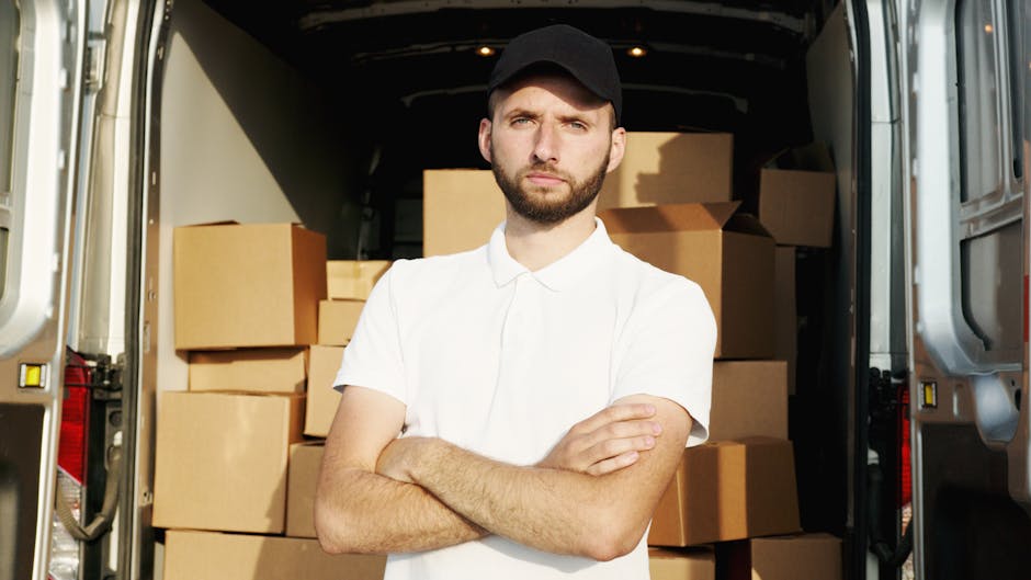 A man with a beard and wearing a red cap, a checkered shirt, and a beige and brown jacket stands inside a van during a house removal process. The interior of the van contains several cardboard boxes,