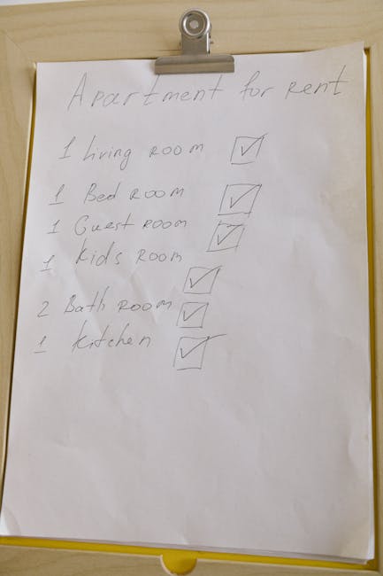A handwritten house relocation checklist on a large sheet of white paper attached to a wall with a metal clip, listing rooms for an apartment rental. The list includes '1 living room,' '1 bed room,' '1 guest room,' '1 kids room,' '2 bath rooms,' and '1 kitchen,' each with a corresponding checked box indicating completion. The paper is slightly curled at the edges, and the background appears to be inside a residential property, possibly near an entryway or moving area. This visual reflects preparation for a home move, relevant to furniture transport and packing tasks undertaken by Man with Van Harmondsworth during a local moving process.
