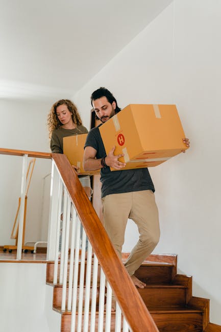 A man and a woman involved in a home relocation are ascending a staircase inside a residential property, carrying cardboard boxes for furniture transport and packing during the moving process. The man, positioned at the front, is holding a large cardboard box with visible tape securing the top, wearing casual clothing and focusing on the task. The woman, following behind, is also carrying a cardboard box, both individual boxes indicating careful packing. The staircase has wooden banisters and steps, with natural indoor lighting illuminating the scene. Behind them, a white wall and a partial view of the upper landing are visible, suggesting an indoor environment used for house moves. Man with Van Harmondsworth, as part of their removals service, manages the logistical aspects of professional furniture transport and packing during home relocations, supporting efficient moving processes.