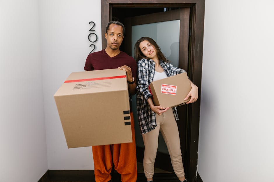 A man with dark hair, wearing an orange traditional garment and a burgundy t-shirt, stands inside a residential doorway holding a large cardboard moving box with black tape and a red 'Fragile' label. Beside him, a woman with long brown hair, dressed in a casual checkered shirt and beige pants, leans against the doorframe while holding a smaller cardboard box with a red 'Fragile' sticker. They are positioned in an indoor entryway, with the door partially open behind them, and are preparing for a home relocation, involving packing and moving furniture and household items. The scene captures the loading process of moving boxes, with visible elements such as packing materials and a neutral interior environment, supporting professional removals services by [COMPANY_NAME].