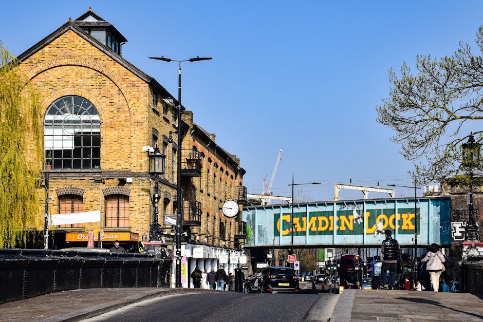 A street scene in an urban area featuring a large, multi-story brick building with arched windows on the upper floor, situated alongside a railway bridge painted in blue with yellow and green lettering reading 'Calden Lock'. The street is busy with pedestrians walking on the sidewalk, some near parked cars, and others approaching the building. Several black vehicles, including a van associated with house removals, are parked along the road, with one van possibly involved in a home relocation or furniture transport process. The scene is illuminated by daylight, and there are street lamps, a clock, and trees with sparse leaves visible on the right side, indicating early spring or late autumn. In the background, cranes and construction equipment suggest ongoing urban development, aligning with the context of packing and moving logistics that Man with Van Harmondsworth might provide during local or long-distance removals.