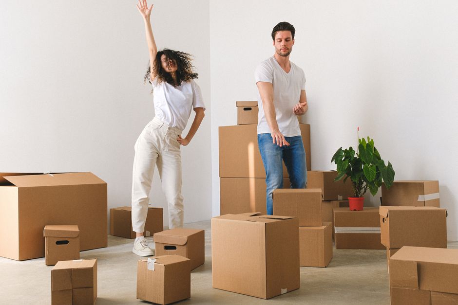 A man and woman are inside a minimally furnished room with white walls during the packing and loading process of a house move. The woman, with curly hair, is standing and raising one arm, wearing casual white clothing and sneakers, while the man, with short dark hair, is wearing a white t-shirt and blue jeans, and is in the act of pointing or reaching towards the packed boxes. Several cardboard boxes of various sizes, some sealed with packing tape, are positioned on the floor around them, with a few stacked against the wall. A small potted plant with green leaves in a red pot is situated on top of a few boxes near the right side of the image. The scene is illuminated by natural light, creating a clear view of the packing and moving materials, which include boxes and a trolley for transporting items. This setup reflects the early stages of a home relocation, with packing supplies, furniture removal, and estate clearing evident, likely managed by a professional removals service such as Man with Van Harmondsworth.
