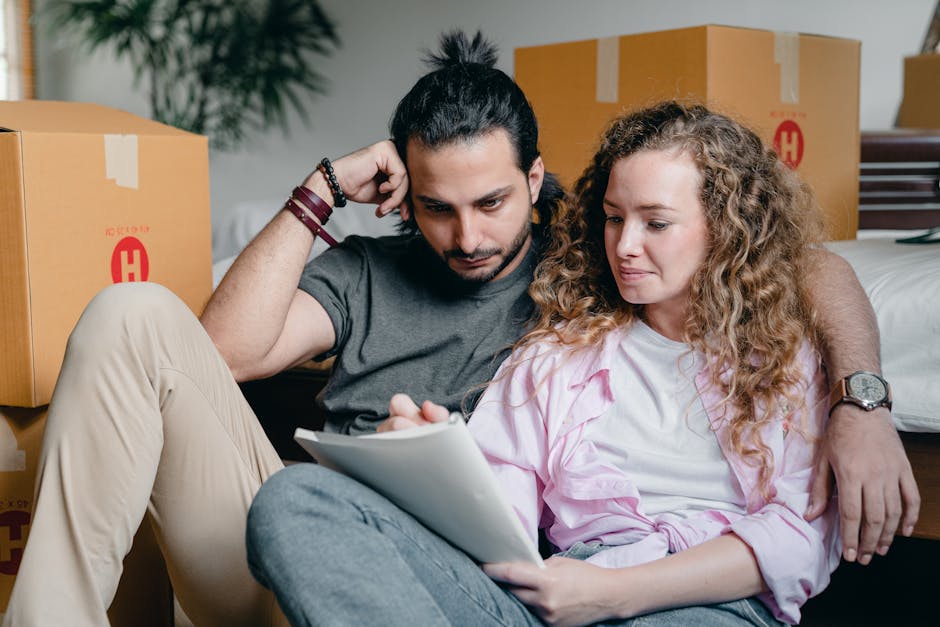 A young couple sitting on the floor inside a room during a house relocation, surrounded by multiple large cardboard boxes with red and yellow markings, some sealed with packing tape. The man, with dark hair tied back and wearing a grey t-shirt, is leaning against the bed frame with his left arm resting on a box, while holding a tablet in his right hand and appearing focused. The woman, with curly blonde hair and wearing a pink and white blouse, is sitting close to him with the tablet on her lap, both looking at the screen with a neutral expression. The room is well-lit, with the bed partly visible behind them, and boxes piled behind and around them indicating an ongoing packing or unpacking process, typical of furniture transport and home packing activities, related to home relocation services offered by Man with Van Harmondsworth.