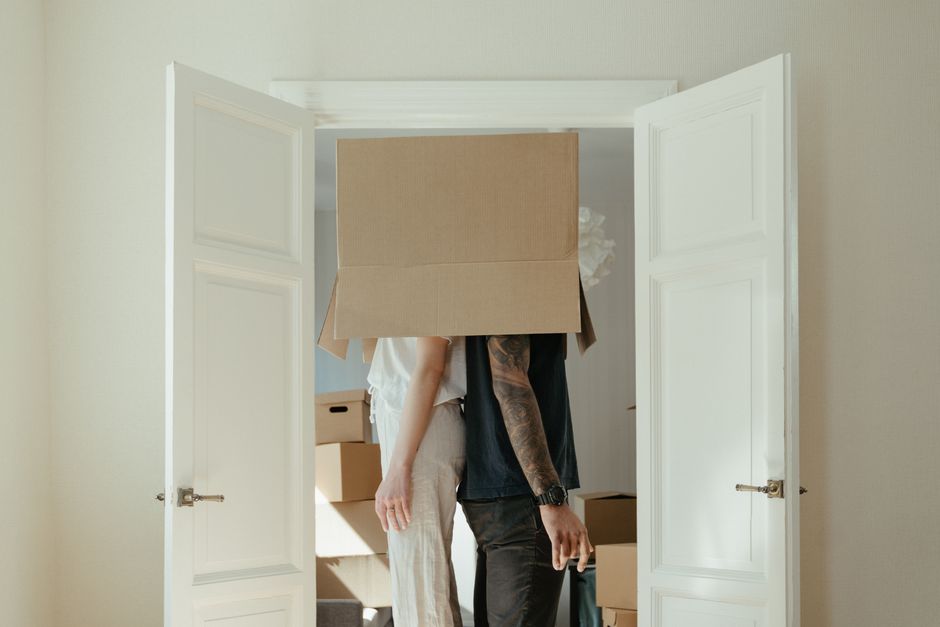A man and woman are inside a minimally furnished room with white walls during the packing and loading process of a house move. The woman, with curly hair, is standing and raising one arm, wearing casual white clothing and sneakers, while the man, with short dark hair, is wearing a white t-shirt and blue jeans, and is in the act of pointing or reaching towards the packed boxes. Several cardboard boxes of various sizes, some sealed with packing tape, are positioned on the floor around them, with a few stacked against the wall. A small potted plant with green leaves in a red pot is situated on top of a few boxes near the right side of the image. The scene is illuminated by natural light, creating a clear view of the packing and moving materials, which include boxes and a trolley for transporting items. This setup reflects the early stages of a home relocation, with packing supplies, furniture removal, and estate clearing evident, likely managed by a professional removals service such as Man with Van Harmondsworth.